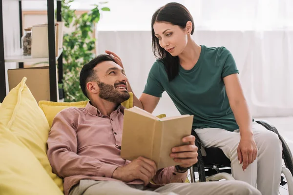 Homme joyeux regardant jolie, petite amie handicapée tout en étant couché sur le canapé et le livre de lecture — Photo de stock