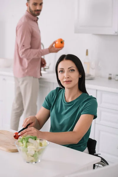 Glückliche behinderte Frau blickt in die Kamera, während sie in der Küche Tomaten in der Nähe ihres Freundes schneidet, der Paprika hält — Stockfoto
