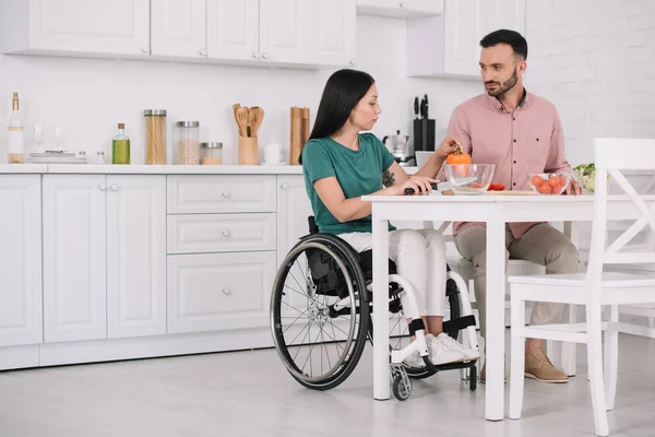 Молодая женщина-инвалид с красивым парнем, сидящая за кухонным столом и готовящая салат вместе — стоковое фото