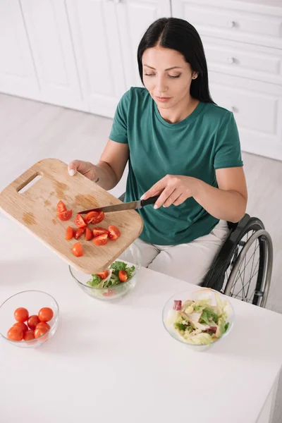 Young disabled woman adding sliced tomatoes in bowl while sitting on wheelchair near kitchen table — Stock Photo