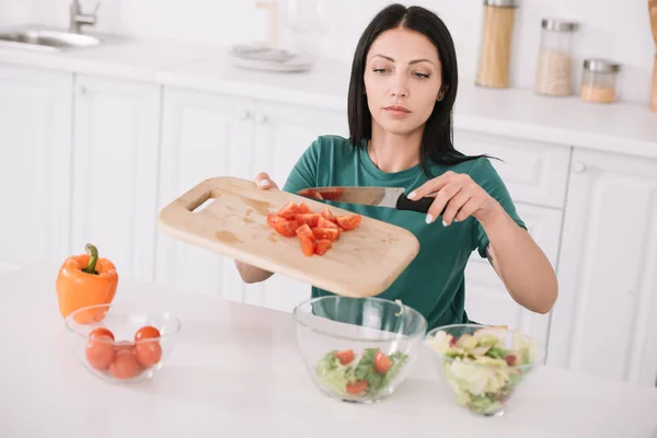 Attractive young woman holding chopping board while adding sliced tomatoes in glass bowl — Stock Photo