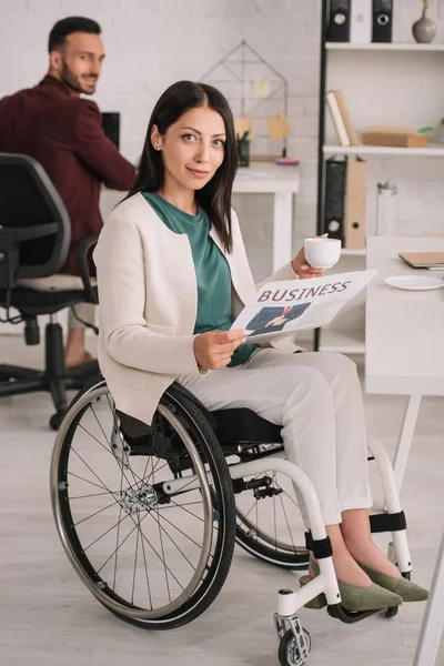 Улыбающаяся деловая женщина-инвалид смотрит в камеру, держа в руках чашку кофе и газету — стоковое фото