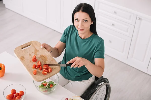 Donna disabile sorridente che guarda la macchina fotografica mentre taglia i pomodori in cucina — Foto stock