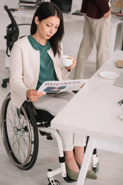 Приваблива бізнес-леді читає газету і тримає чашку кави, сидячи в інвалідному візку на робочому місці — стокове фото