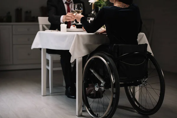 Vista parcial de la joven en silla de ruedas tintineo vasos de vino blanco con novio durante la cena romántica - foto de stock