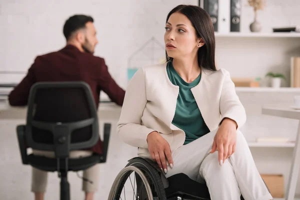 Grave mulher de negócios com deficiência olhando para longe enquanto sentada em cadeira de rodas no escritório perto colega — Fotografia de Stock