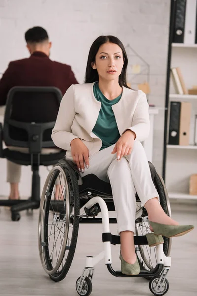 Confident, disabled businesswoman looking at camera while sitting in wheelchair in office near colleague working on background — Stock Photo