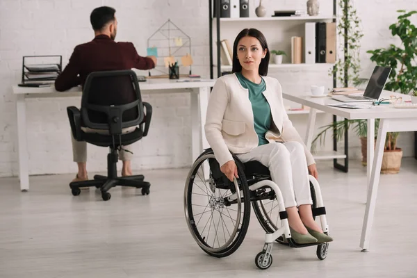 Atractiva mujer de negocios discapacitada sentada en silla de ruedas en oficina cerca de colega - foto de stock