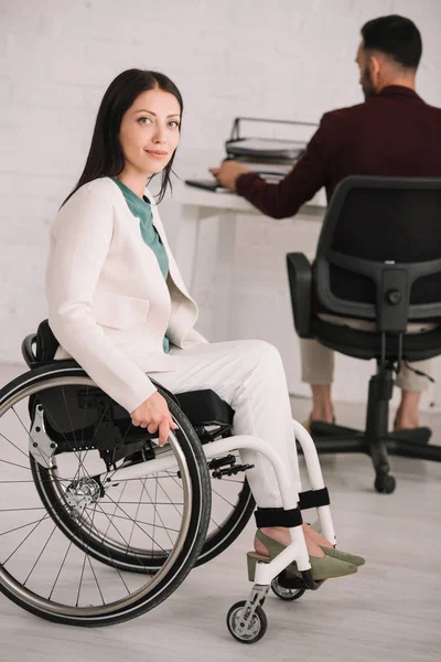 Atraente mulher de negócios deficiente sorrindo para a câmera enquanto sentado em cadeira de rodas perto colega trabalhando no fundo — Fotografia de Stock