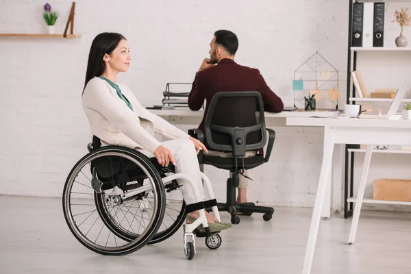 Donna d'affari disabile sorridente che distoglie lo sguardo mentre si siede sulla sedia a rotelle vicino al collega — Foto stock