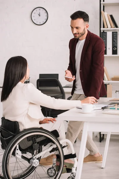 Sonriente hombre de negocios sosteniendo teléfono inteligente mientras está de pie cerca de mujer de negocios discapacitada en la oficina - foto de stock