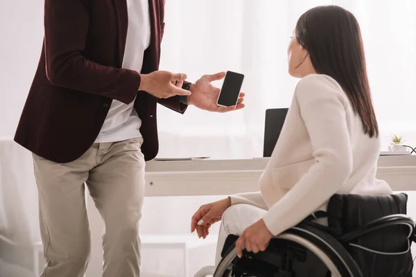 Vista recortada del administrador que muestra el teléfono inteligente con pantalla en blanco a la mujer de negocios con discapacidad - foto de stock