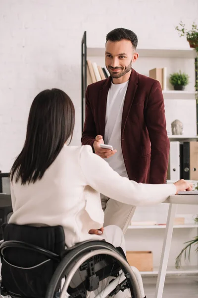 Lächelnder Geschäftsmann steht neben behinderter Geschäftsfrau, während er sein Smartphone im Büro hält — Stockfoto