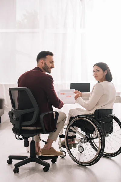 Donna d'affari disabile sorridente che guarda la macchina fotografica mentre si siede vicino al partner d'affari e in possesso di documenti — Foto stock