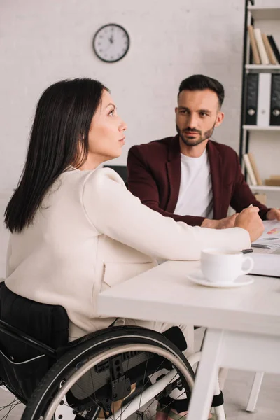 Pensativo discapacitado mujer de negocios sentado en el escritorio junto con apuesto socio de negocios - foto de stock