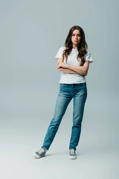 Sad beautiful girl in white t-shirt and jeans with crossed arms isolated on grey — Stock Photo