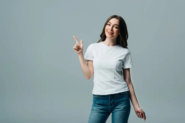 Sonriente hermosa chica en camiseta blanca señalando con el dedo a un lado aislado en gris - foto de stock