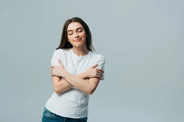 Dreamy smiling beautiful girl in white t-shirt hugging herself with closed eyes isolated on grey — Stock Photo