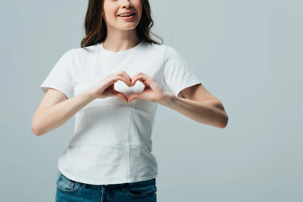 Vista ritagliata di felice bella ragazza in t-shirt bianca mostrando gesto del cuore isolato su grigio — Foto stock