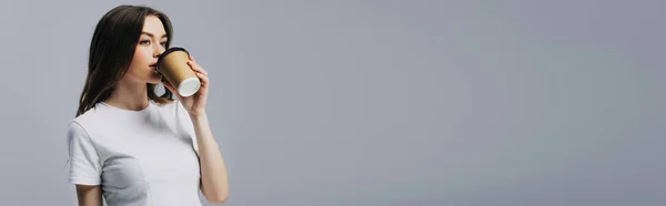 Panoramic shot of beautiful girl in white t-shirt drinking coffee to go isolated on grey — Stock Photo