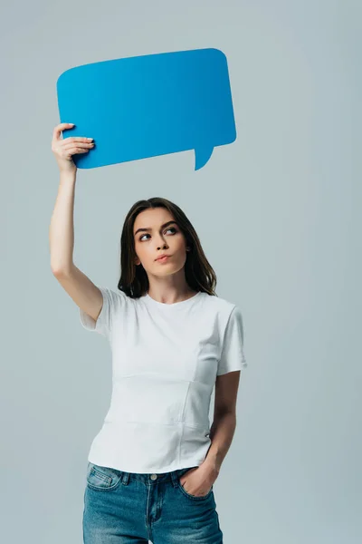 Pensativa hermosa chica en camiseta blanca sosteniendo en blanco burbuja de habla azul aislado en gris - foto de stock