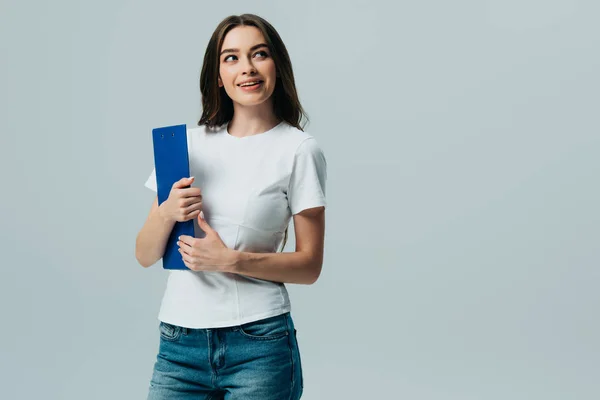 Feliz hermosa chica en blanco camiseta sujetando portapapeles mirando hacia otro lado aislado en gris - foto de stock