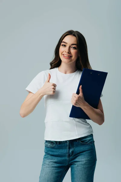 Feliz hermosa chica en blanco camiseta sujetando portapapeles mostrando el pulgar hacia arriba aislado en gris - foto de stock