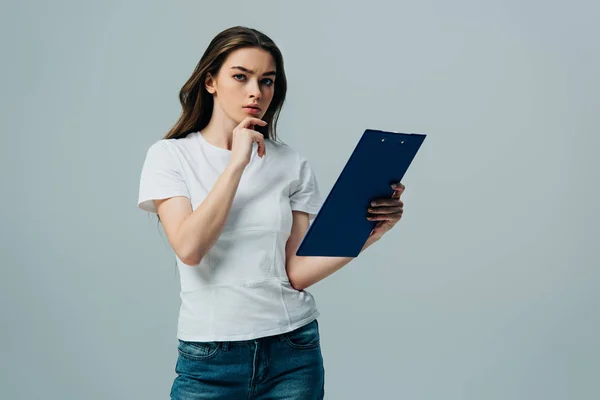 Pensive beautiful girl in white t-shirt holding blue clipboard isolated on grey — Stock Photo