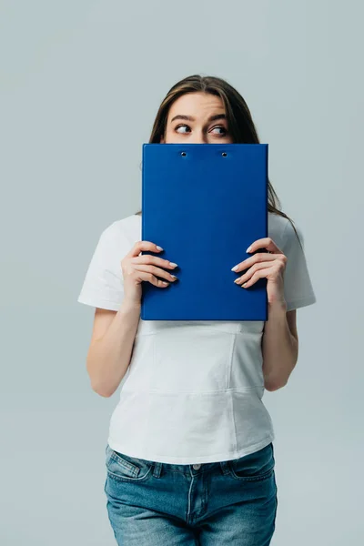 Beautiful girl in white t-shirt holding blue clipboard in front of face and looking away isolated on grey — Stock Photo