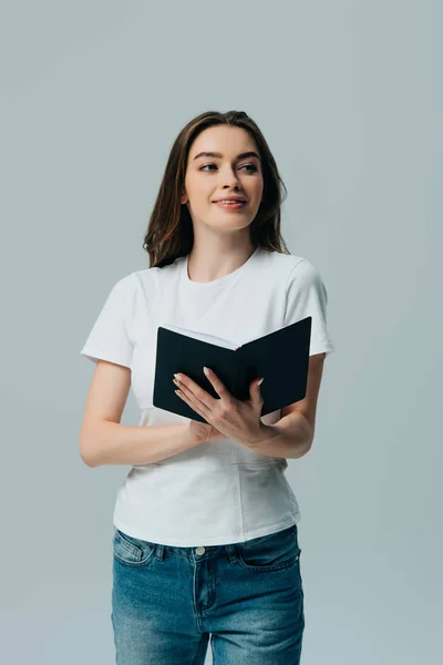 Happy beautiful girl in white t-shirt holding notebook and looking away isolated on grey — Stock Photo