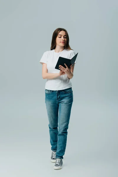 Красива дівчина в білій футболці і джинсах читає книгу ізольовано на сірому — стокове фото