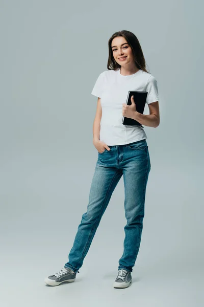 Счастливая красивая девушка в белой футболке и джинсах с блокнотом изолированы на сером — стоковое фото