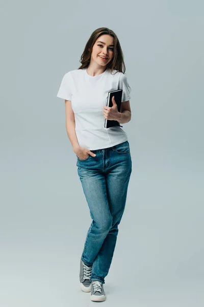 Щаслива красива дівчина в білій футболці і джинсах тримає блокнот ізольовано на сірому — стокове фото