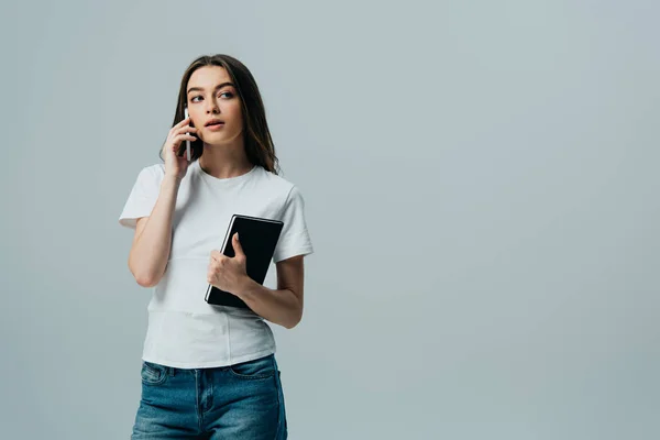 Мрійлива красива дівчина в білій футболці говорить на смартфоні і тримає блокнот ізольовано на сірому — стокове фото