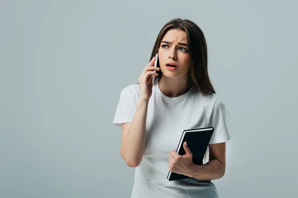 Sad and surprised beautiful girl in white t-shirt talking on smartphone and holding notebook isolated on grey — Stock Photo