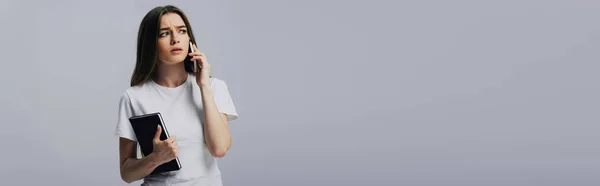 Triste bella ragazza in t-shirt bianca parlando su smartphone e tenendo il taccuino isolato su grigio, colpo panoramico — Foto stock