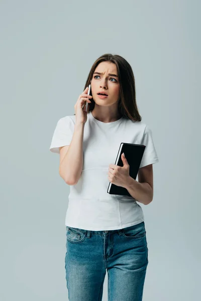 Shocked beautiful girl in white t-shirt talking on smartphone and holding notebook isolated on grey — Stock Photo