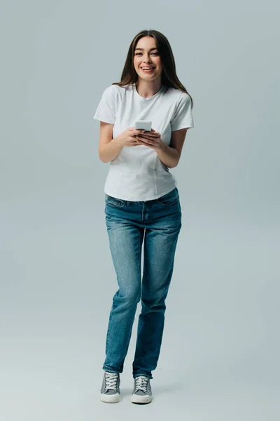 Happy beautiful girl in white t-shirt and jeans using smartphone isolated on grey — Stock Photo
