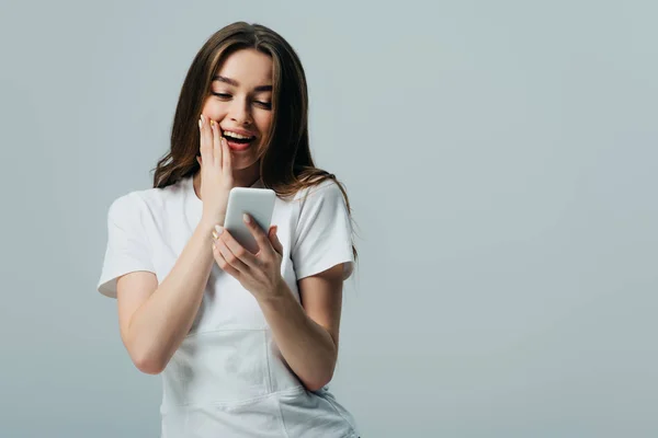 Emocionado impactado hermosa chica en camiseta blanca usando teléfono inteligente aislado en gris - foto de stock