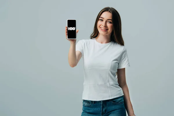 KYIV, UCRANIA - 6 de junio de 2019: hermosa chica feliz en camiseta blanca que muestra el teléfono inteligente con la aplicación HBO aislada en gris - foto de stock