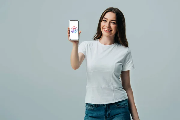 KYIV, UKRAINE - JUNHO 6, 2019: menina bonita feliz em camiseta branca mostrando smartphone com ícone de música de maçã isolado em cinza — Fotografia de Stock