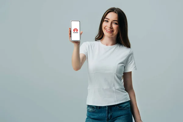 KYIV, UCRANIA - 6 DE JUNIO DE 2019: hermosa niña feliz en camiseta blanca que muestra el teléfono inteligente con el logotipo de hue.net aislado en gris - foto de stock