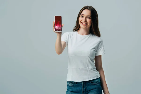 Menina bonita feliz em t-shirt branca mostrando smartphone com cursos de negociação aplicativo isolado em cinza — Fotografia de Stock