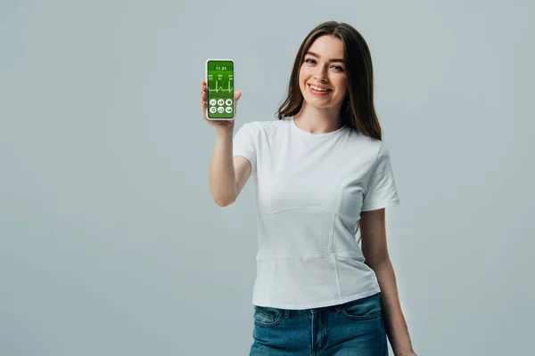 Happy beautiful girl in white t-shirt showing smartphone with healthcare app isolated on grey — Stock Photo