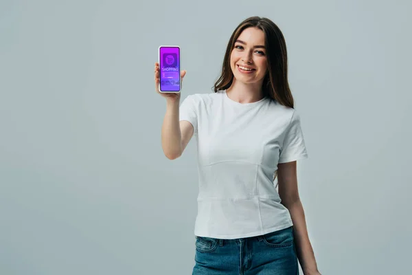 Feliz hermosa chica en camiseta blanca que muestra el teléfono inteligente con aplicación de compras en línea aislado en gris - foto de stock