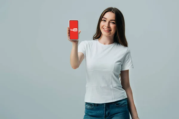 KYIV, UCRANIA - 6 de junio de 2019: hermosa chica feliz en camiseta blanca que muestra el teléfono inteligente con la aplicación de youtube aislada en gris - foto de stock