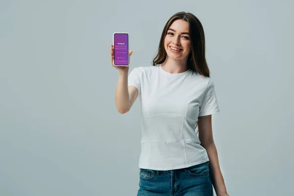 KYIV, UCRANIA - 6 de junio de 2019: hermosa chica feliz en camiseta blanca que muestra el teléfono inteligente con la aplicación de Instagram aislada en gris - foto de stock