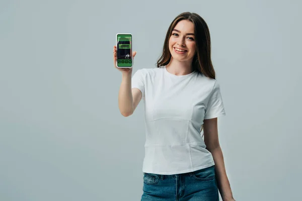 Счастливая красивая девушка в белой футболке показывая смартфон с приложением бронирования изолированы на серый — стоковое фото