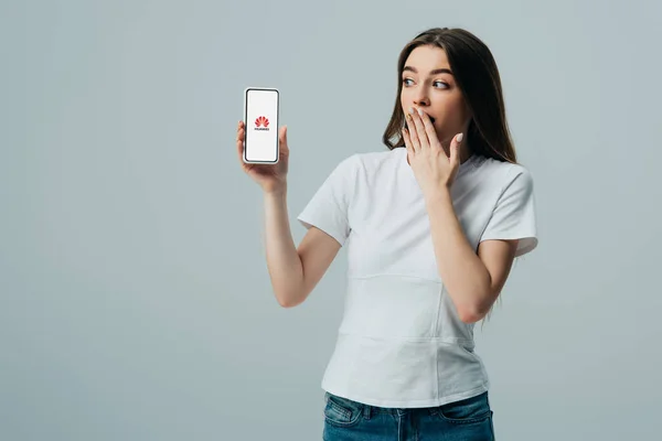 KYIV, UCRANIA - 6 DE JUNIO DE 2019: impactada hermosa chica en camiseta blanca que muestra el teléfono inteligente con el logotipo de hue.net aislado en gris - foto de stock