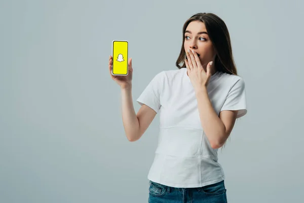 KYIV, UCRAINA - 6 GIUGNO 2019: stupita bella ragazza in t-shirt bianca che mostra smartphone con app Snapchat isolata su grigio — Foto stock